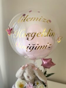 Yakın çekim: şeffaf balonda altın "Ailemize Hoşgeldin Miniğimiz" yazısı, içinde pembe balonlar, konfeti ve pembe kelebekler