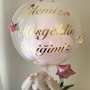 Yakın çekim: şeffaf balonda altın "Ailemize Hoşgeldin Miniğimiz" yazısı, içinde pembe balonlar, konfeti ve pembe kelebekler