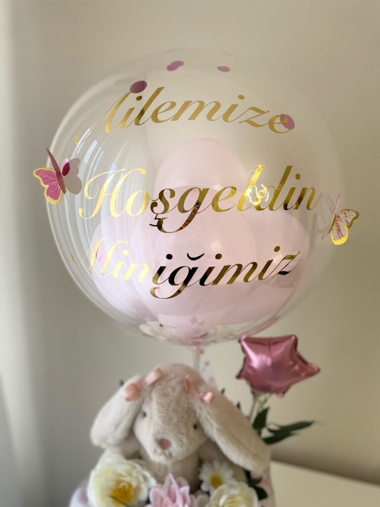 Yakın çekim: şeffaf balonda altın "Ailemize Hoşgeldin Miniğimiz" yazısı, içinde pembe balonlar, konfeti ve pembe kelebekler
