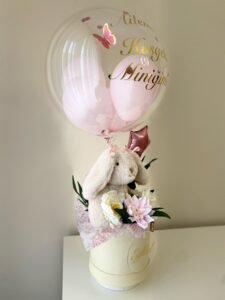 Pembe iç balonlu şeffaf bobo balonun altında peluş tavşan ve çiçekler, krem kutu yan görünümü; yenidoğan balon kutusu