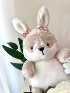 Yakın çekim: kulaklarında pembe fiyonk olan bej-beyaz peluş tavşan, yanında yeşil yapraklar ve beyaz çiçek