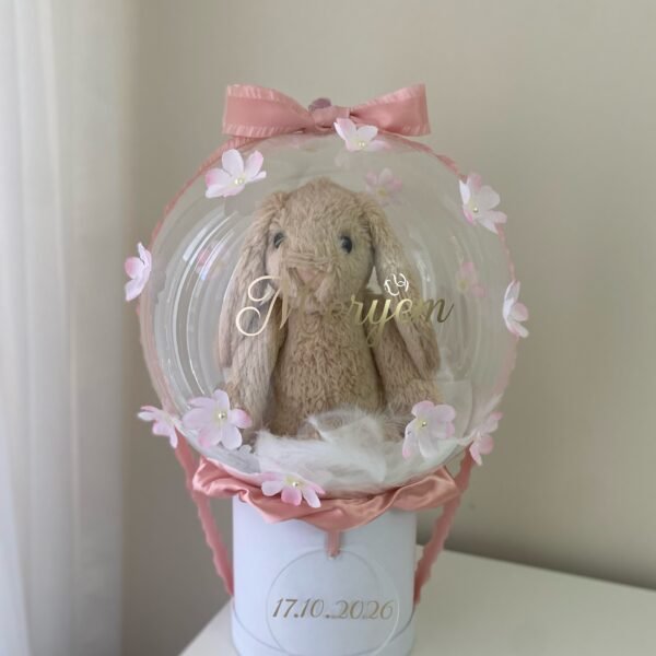 Şeffaf balon içinde tüyler üstünde peluş tavşan, altın isim yazısı, incili pembe çiçekler, beyaz kutu; kız bebek balon kutusu