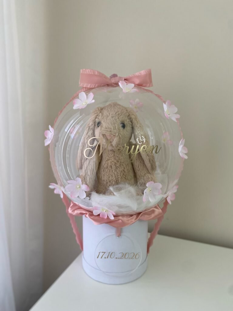 Şeffaf balon içinde tüyler üstünde peluş tavşan, altın isim yazısı, incili pembe çiçekler, beyaz kutu; kız bebek balon kutusu