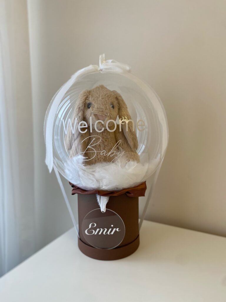 Beyaz "Welcome Baby" yazılı şeffaf balon içinde peluş tavşan ve tüyler, kahverengi kutu ve isimli pleksi; baby balon kutusu