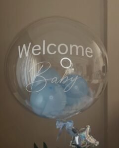 Yakın çekim: şeffaf balondaki beyaz "Welcome Baby" yazısının ardında açık mavi balonlar ve beyaz tüyler