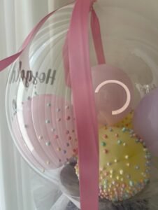 Yakın çekim: şeffaf balonun içinde pastel köpük toplarla kaplı sarı ve pembe balonlar, önünde pembe saten kurdele